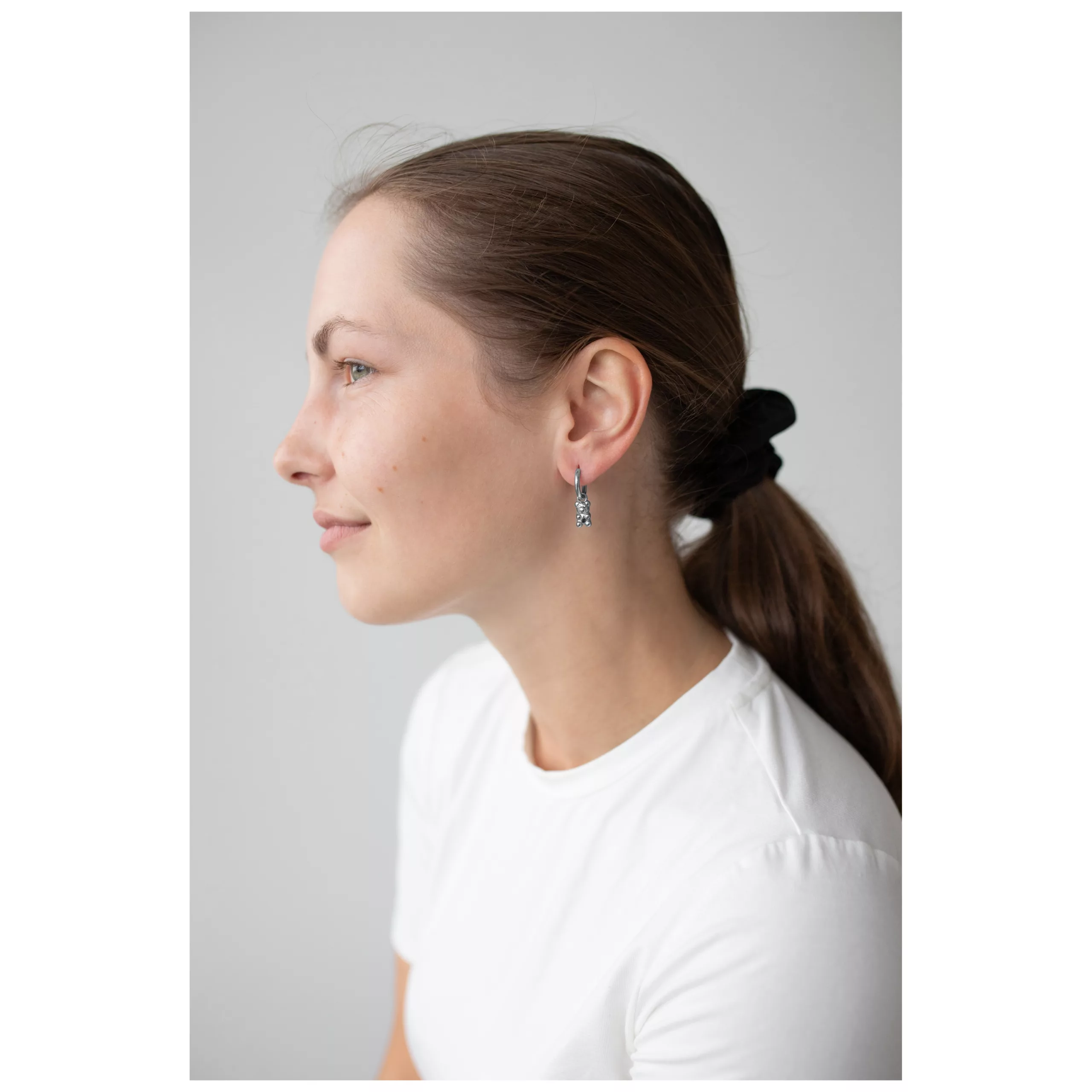 Donna con capelli castani indossa orecchini a cerchio con orsetti gommosi neri, maglietta bianca.