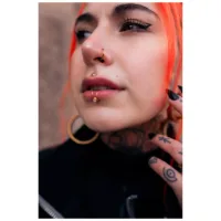 Giovane persona con capelli arancioni indossa un lipcuff argento e orecchini dorati su sfondo neutro
