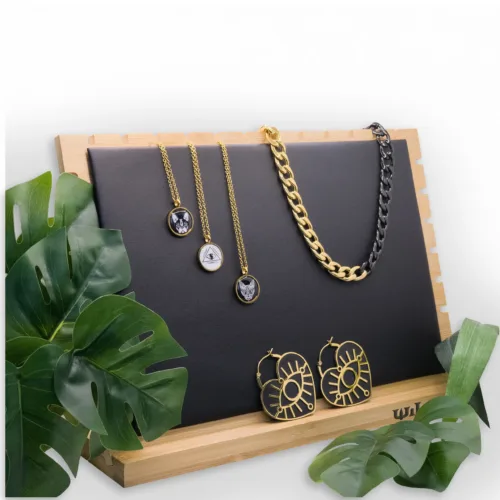 Espositore nero per gioielli con catene dorate e orecchini su cornice in legno, circondato da foglie verdi