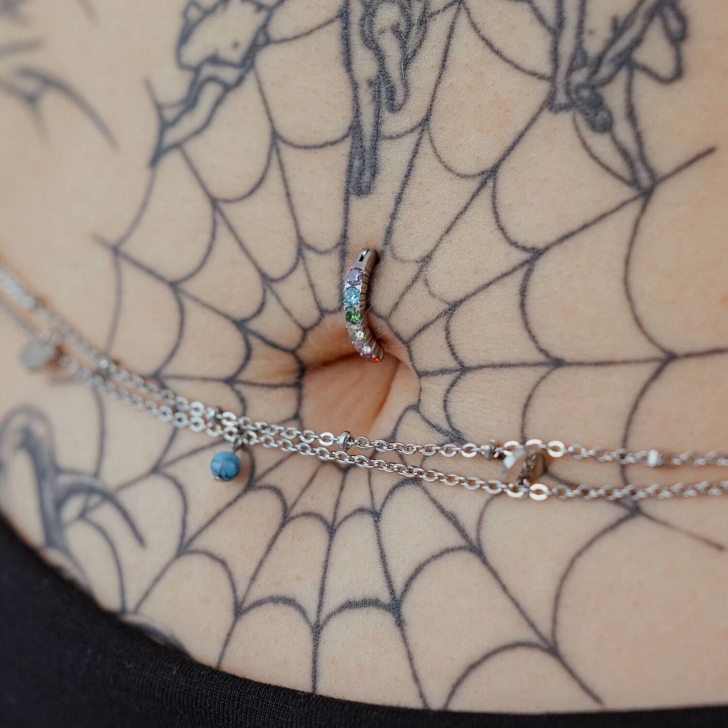 Piercing all'ombelico con cristalli arcobaleno su pelle tatuata, primo piano