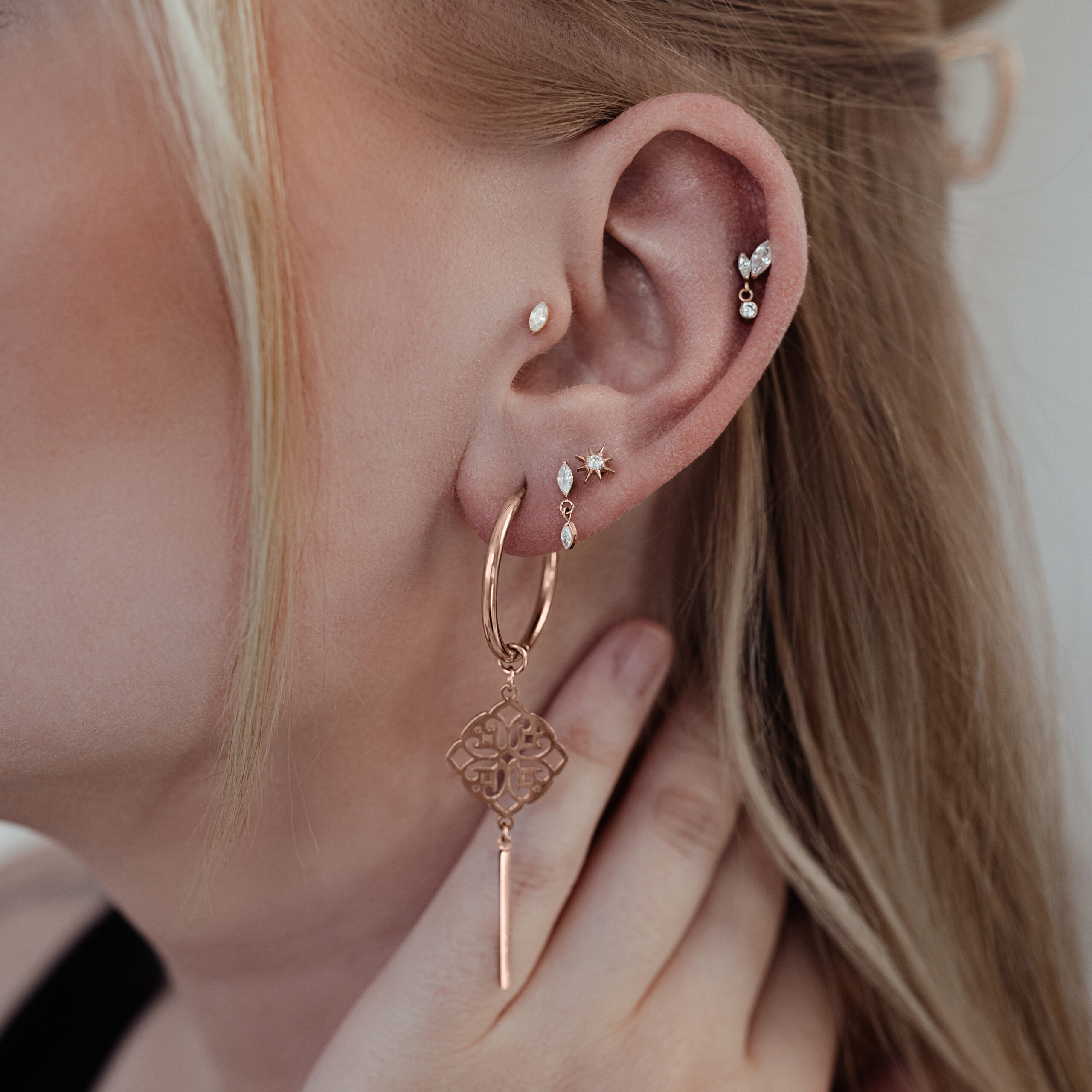 Orecchio con diversi piercing, incluso un grande ciondolo ovale in cristallo oro rosa su sfondo chiaro