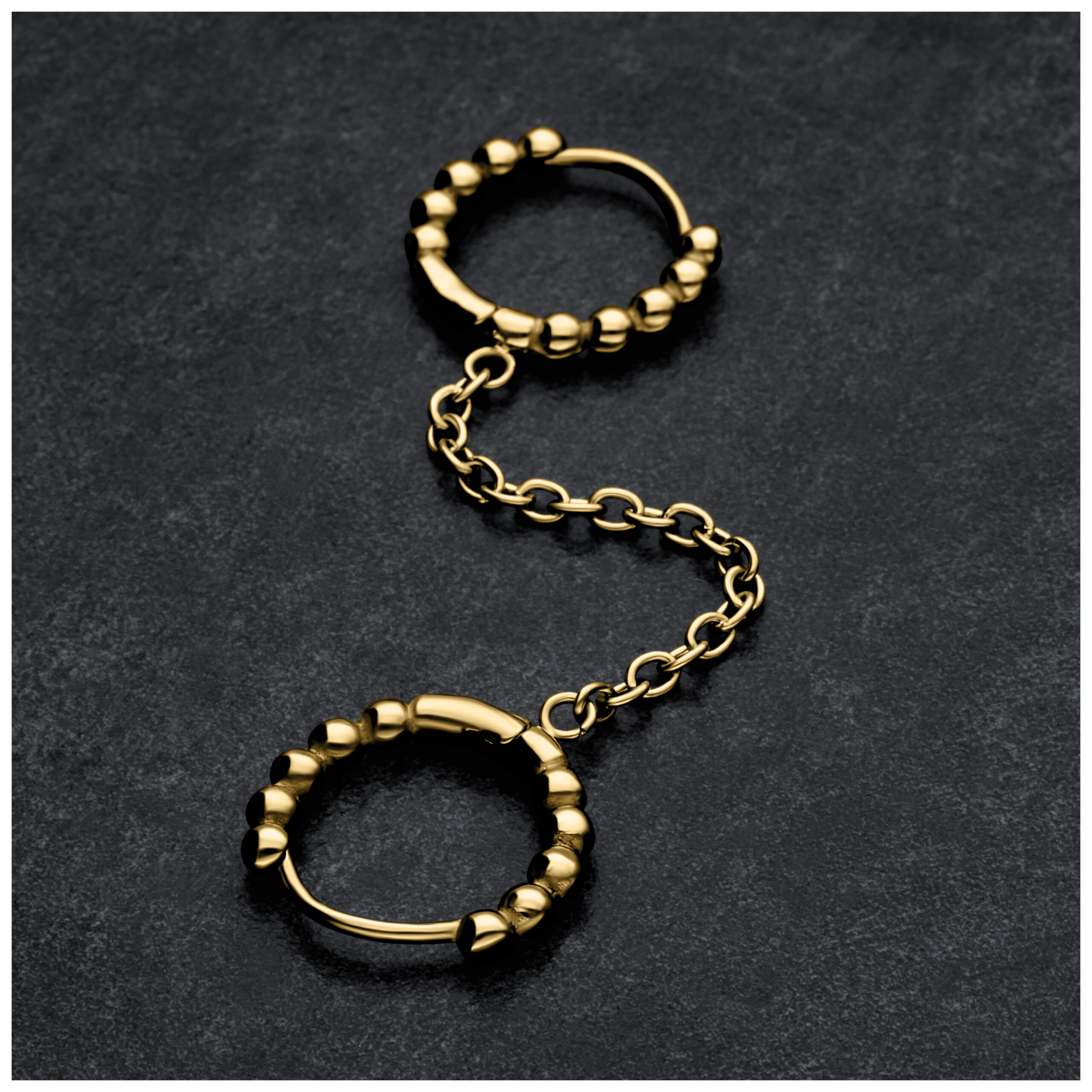 Doppelte Huggie-Ohrpiercings in Gold mit Kugeln und Kette auf dunklem Hintergrund