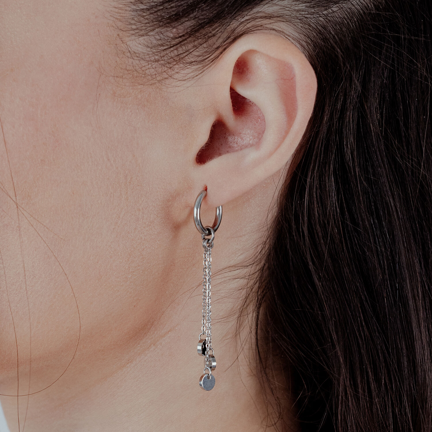 Orecchini a cerchio argentati con pendente a catena su orecchio femminile su sfondo chiaro
