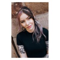 Donna con orecchini neri e argento e piercing, sorridente davanti a un muro di pietra