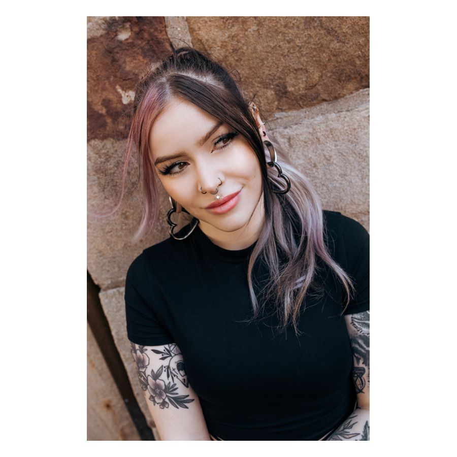 Donna con orecchini neri e argento e piercing, sorridente davanti a un muro di pietra