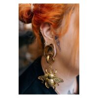 Donna con capelli rosso-arancio indossa orecchino dorato con grande ciondolo ape su sfondo sfocato