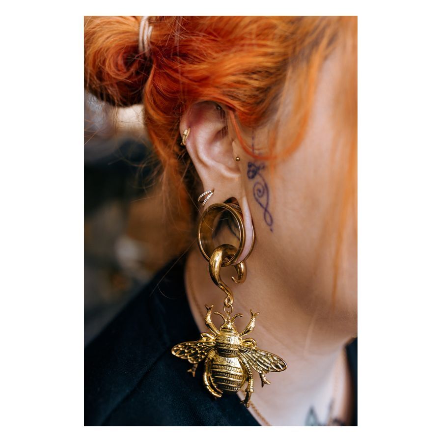 Donna con capelli rosso-arancio indossa orecchino dorato con grande ciondolo ape su sfondo sfocato