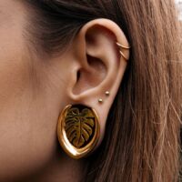 Orecchini Monstera dorati, indossati nell'orecchio, con piercing sullo sfondo