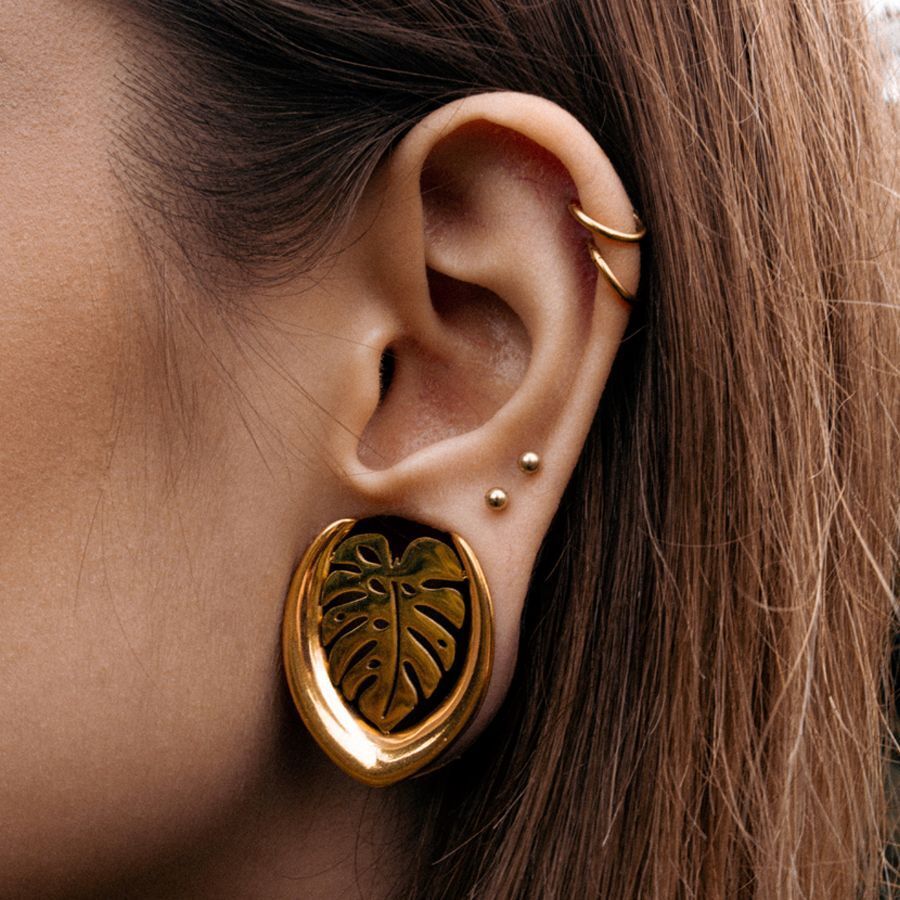 Orecchini Monstera dorati, indossati nell'orecchio, con piercing sullo sfondo