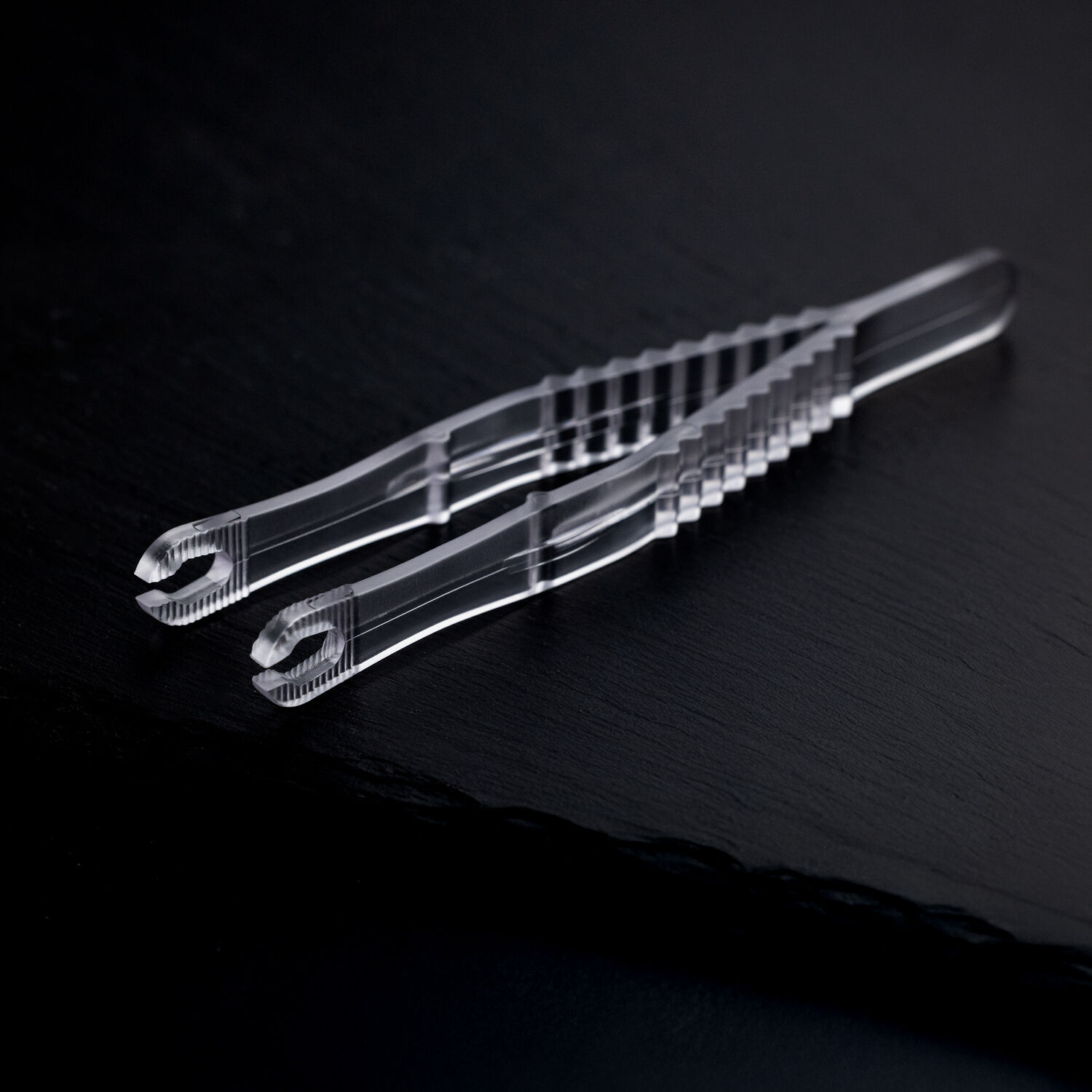 Due pinze da piercing rotonde e sterili trasparenti incrociate su uno sfondo scuro.