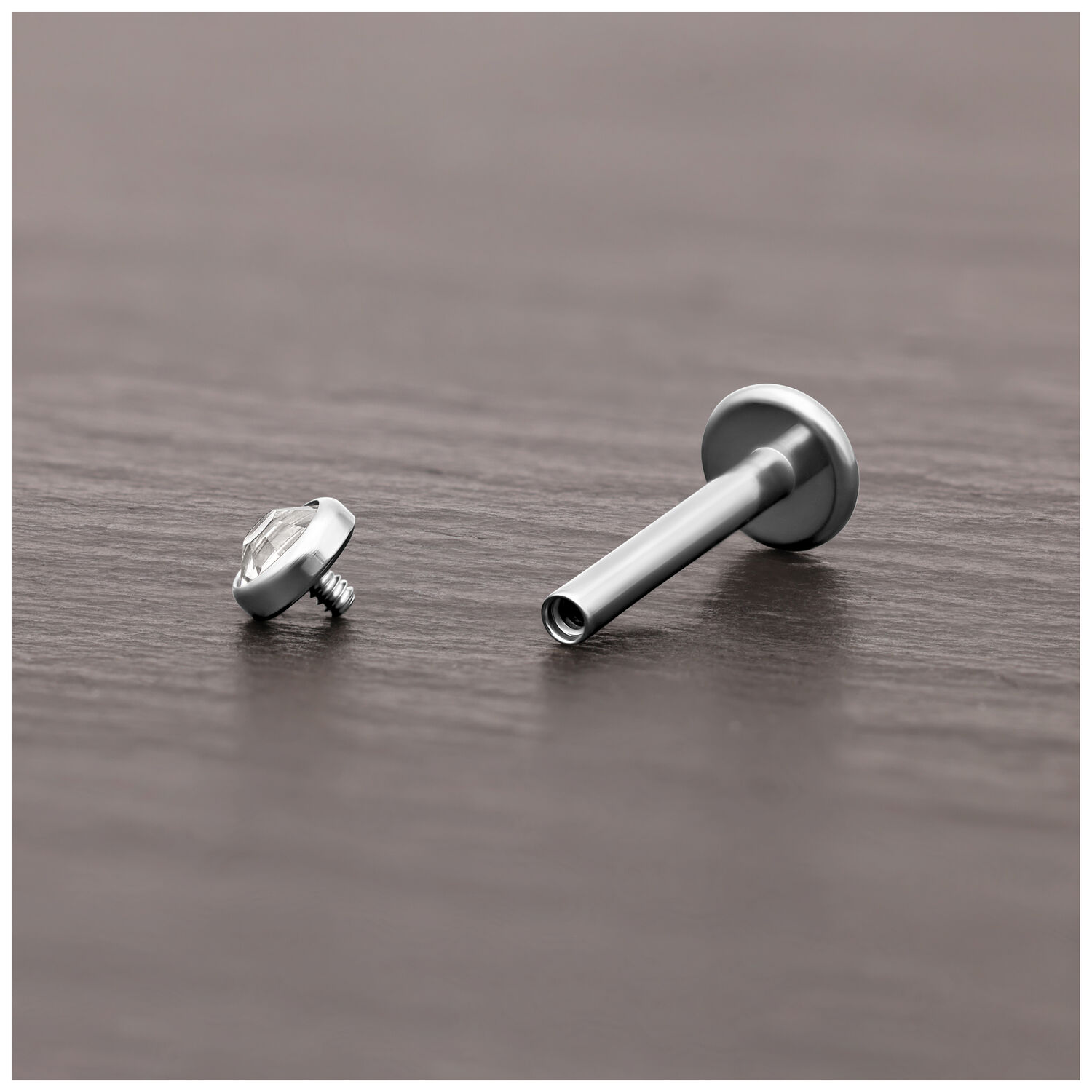 Attacco per piercing in titanio argento con cristallo chiaro su legno scuro.