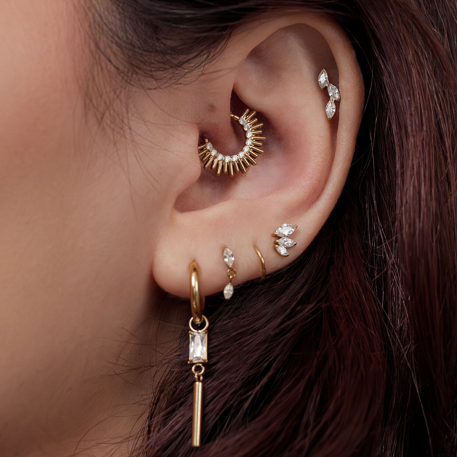 Orecchio con diversi piercing dorati, incluso un Sun Beams Clicker, davanti a capelli castani