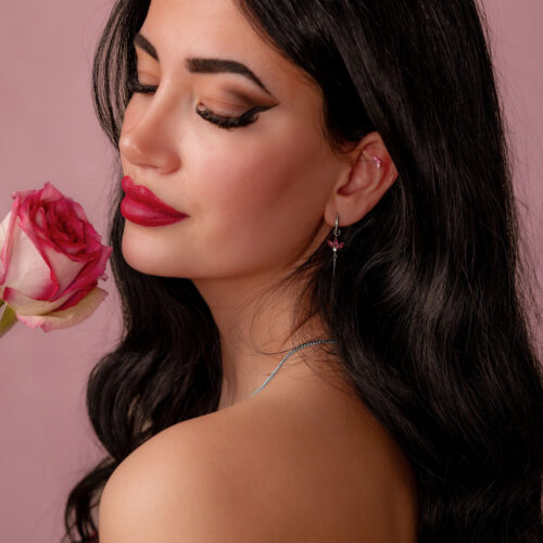Donna con lunghi capelli scuri tiene una rosa rosa e indossa un orecchino argento con cristallo rosso su sfondo rosa