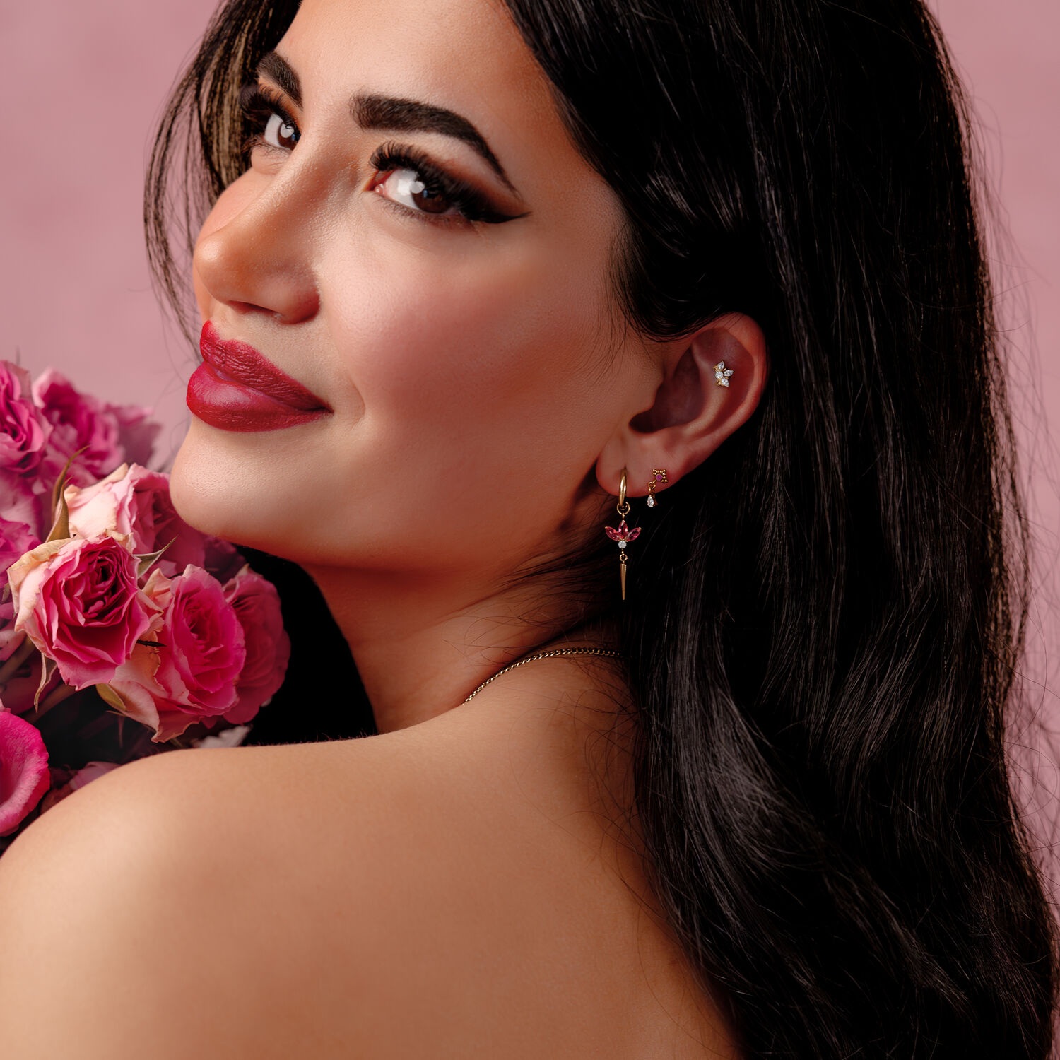 Donna con lunghi capelli neri indossa orecchini d'argento con cristalli rossi, tiene rose rosa su sfondo rosa