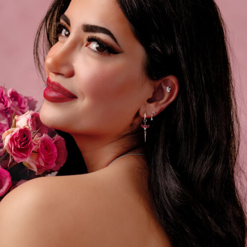 Donna con lunghi capelli scuri indossa orecchini d'argento con cristalli rossi e tiene rose rosa su sfondo rosa.