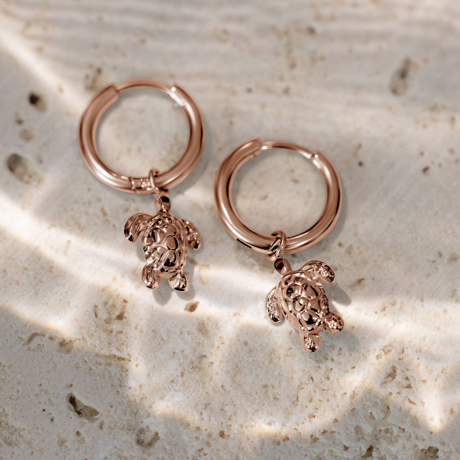 Orecchini a cerchio in oro rosa con piccoli ciondoli a tartaruga su superficie chiara e strutturata