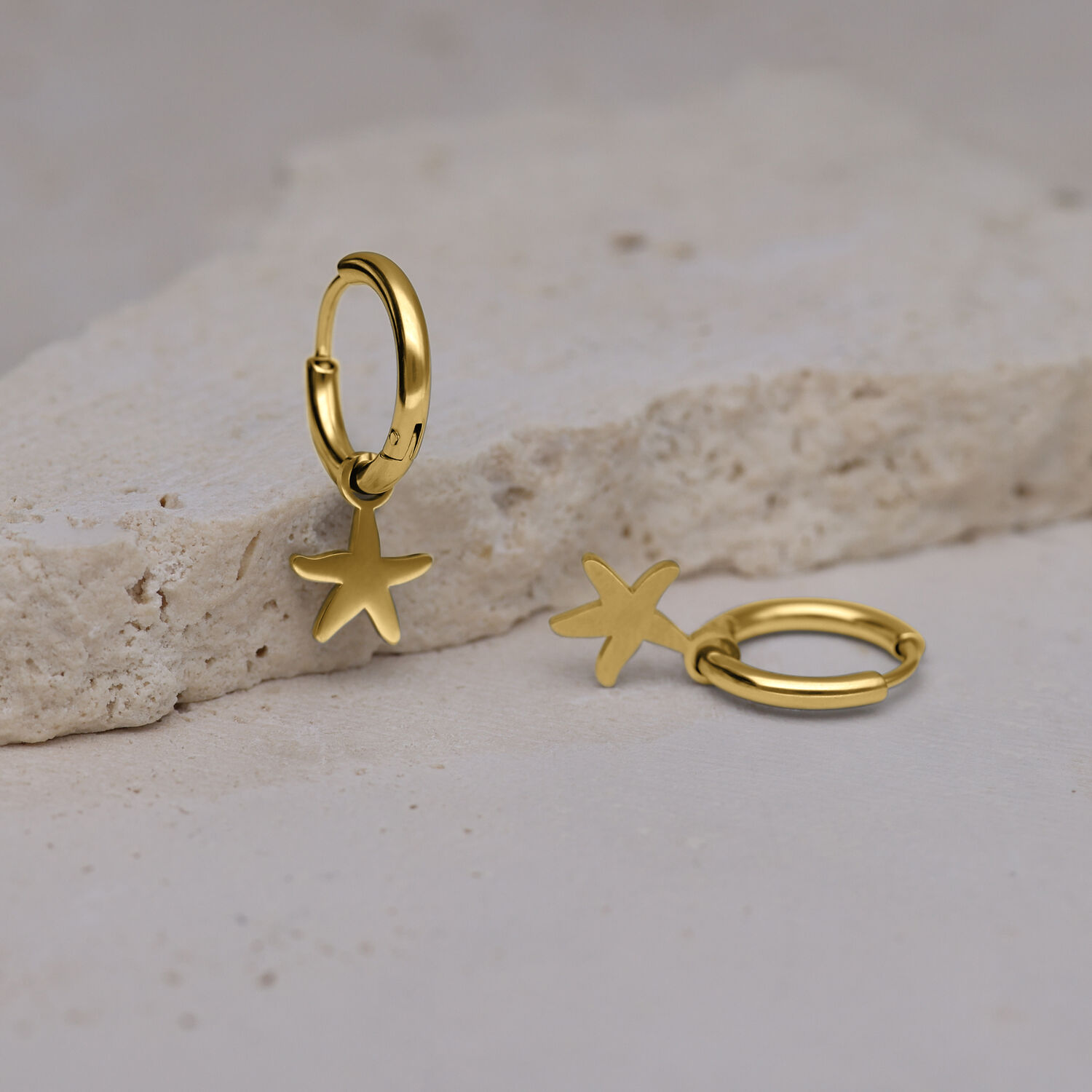 Mini Orecchini a Cerchio Zlata con Stelle Marine Dorate Due orecchini a cerchio dorati con ciondoli a stella marina su una superficie di pietra