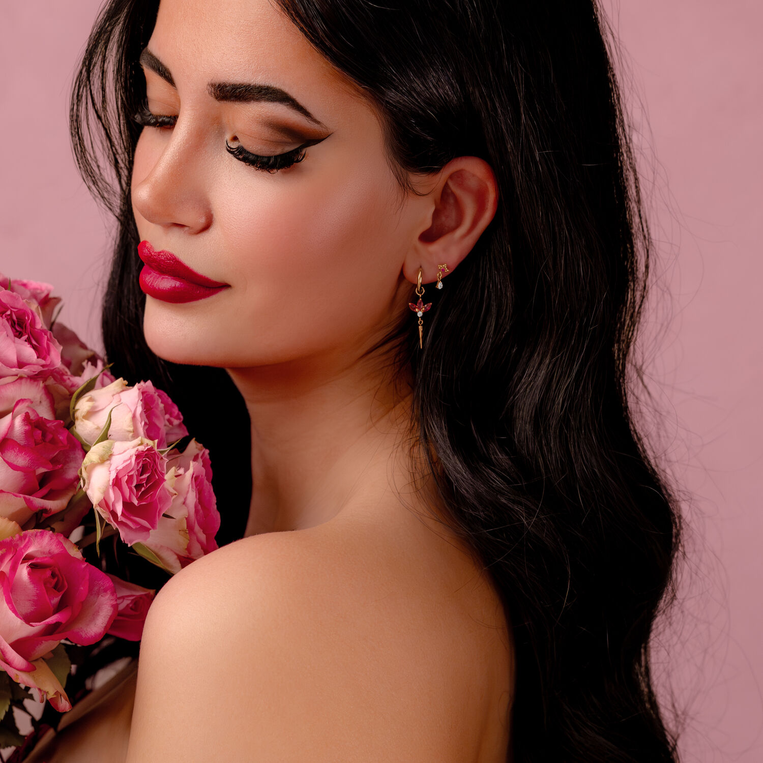 Donna con lunghi capelli neri, labbra rosse e orecchini dorati, tiene rose rosa su sfondo rosa