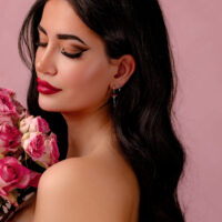 Donna con lunghi capelli scuri e orecchini d'argento con cristalli rossi, tiene fiori rosa su sfondo rosa