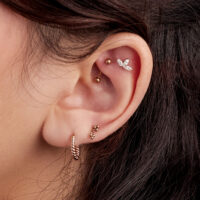 Orecchio con più piercing, tra cui un motivo a foglia in oro rosa e un orecchino a cerchio intrecciato.