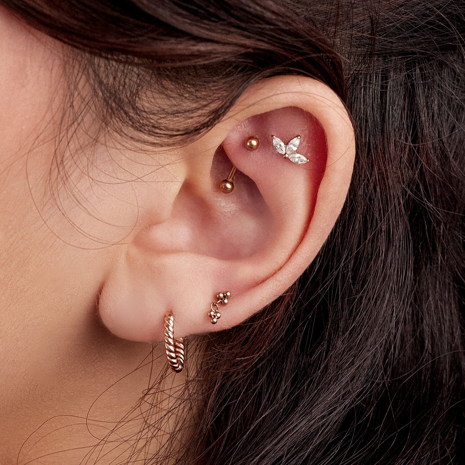 Orecchio con più piercing, tra cui un motivo a foglia in oro rosa e un orecchino a cerchio intrecciato.