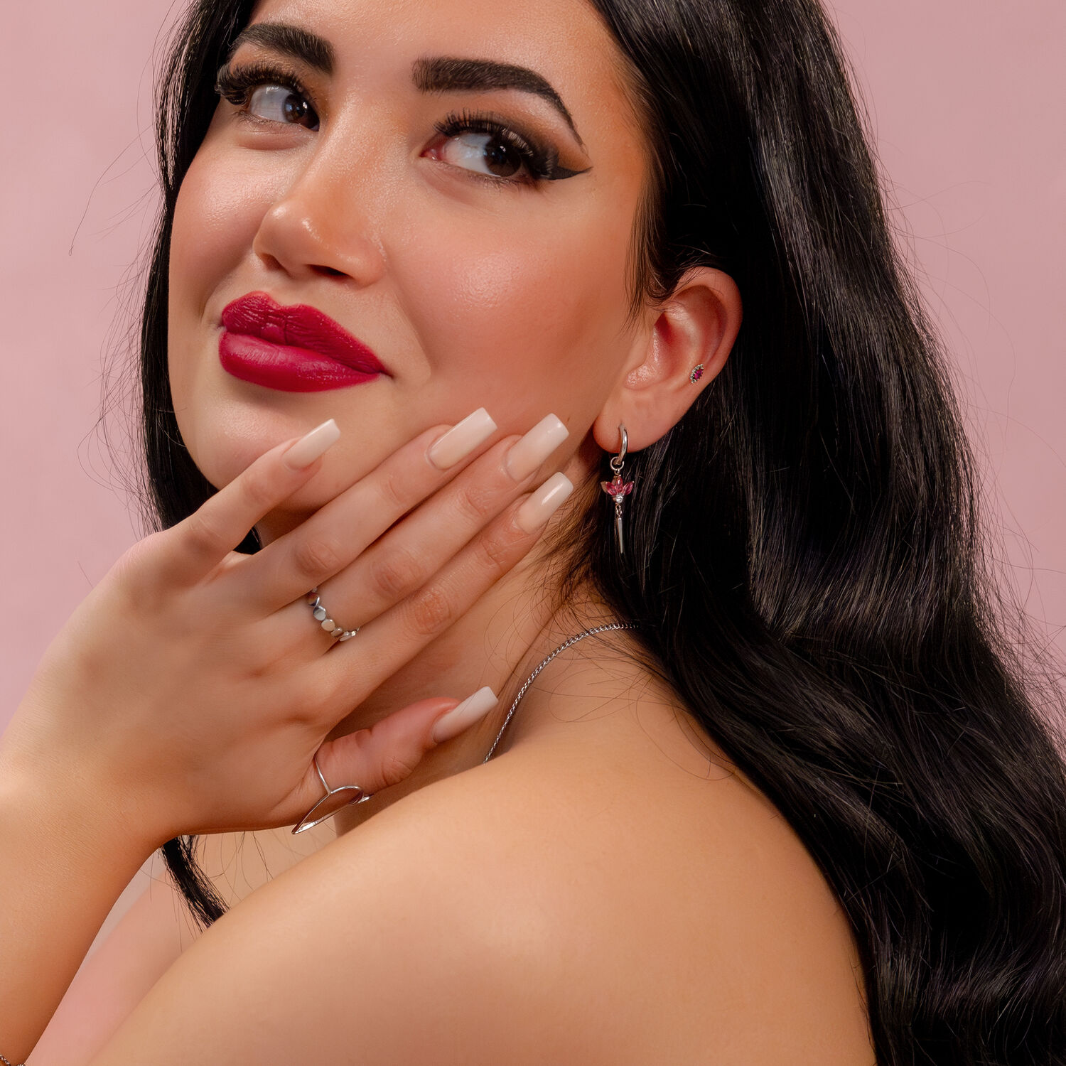 Donna con lunghi capelli neri, labbra rosse e orecchino d'argento con cristallo rosso su sfondo rosa
