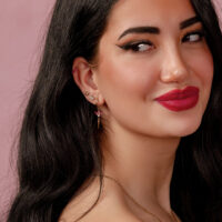 Donna con capelli neri che indossa diversi orecchini, incluso un attacco botanico con rubino su sfondo rosa.