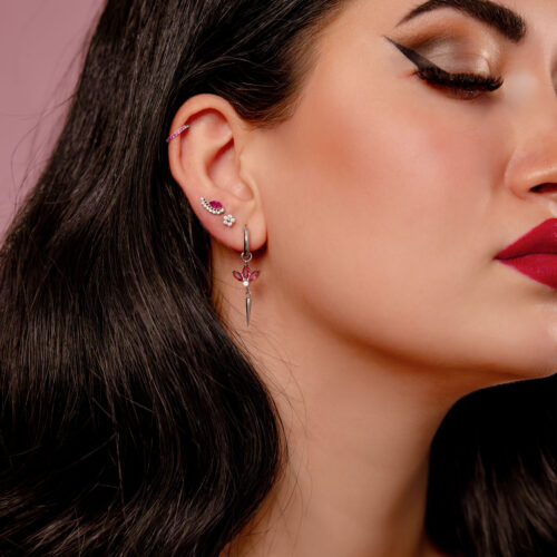 Primo piano di un orecchio con più piercing, incluso un clicker in argento con cristalli rossi, su sfondo rosa.