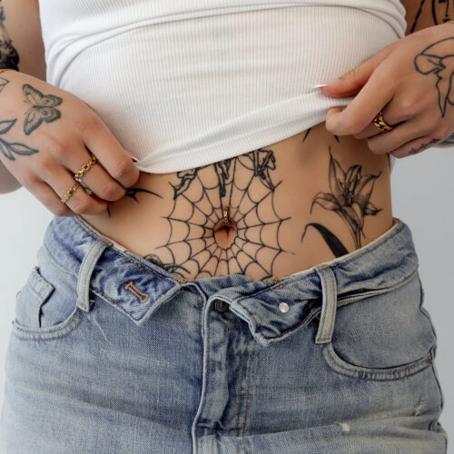 Piercing all'ombelico con motivo leopardato dorato, indossato da persona con tatuaggi e jeans.