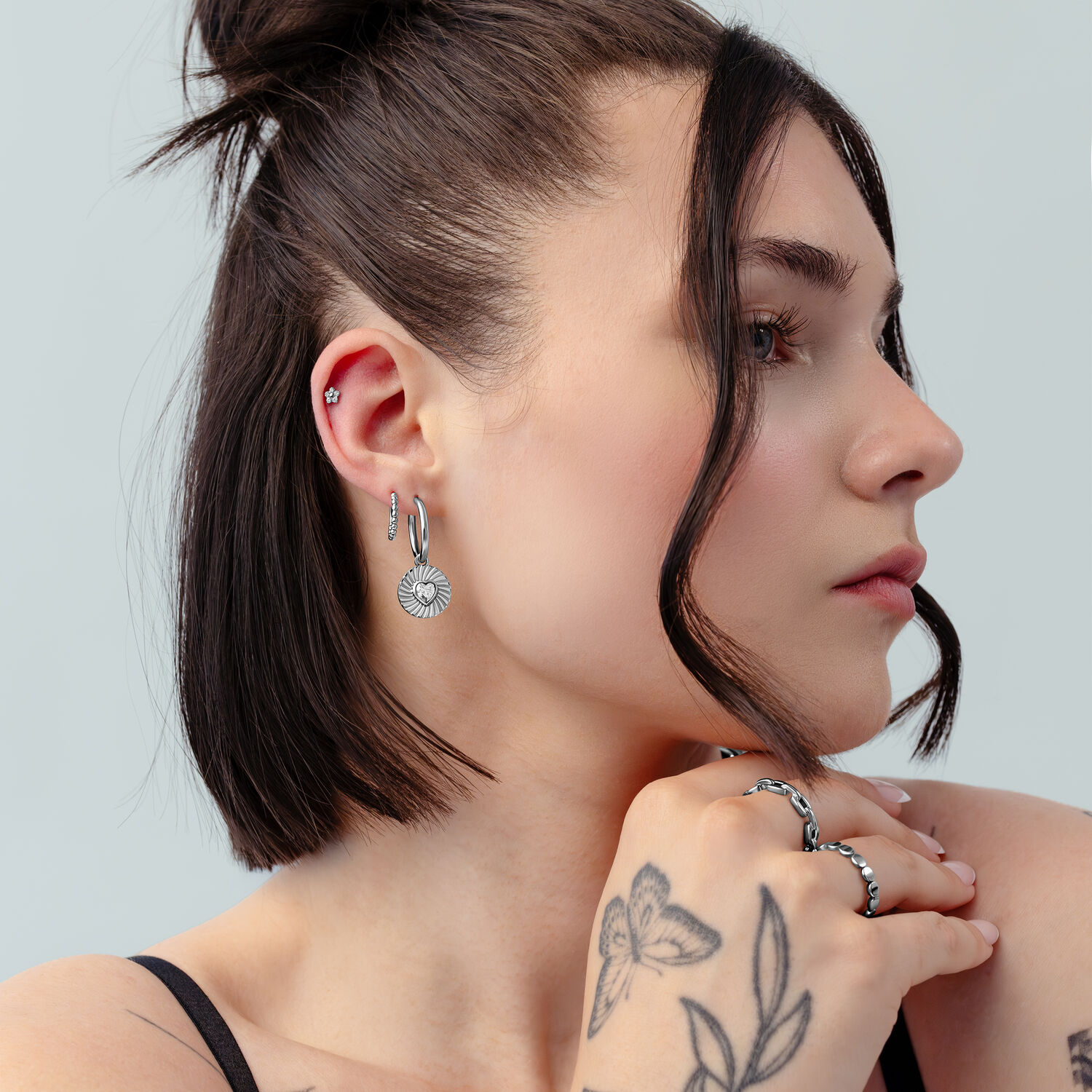 Creole a Cuore Riffled Argento in Profilo Donna con capelli corti e scuri indossa creole argento con design a cuore e tatuaggi su sfondo grigio chiaro.