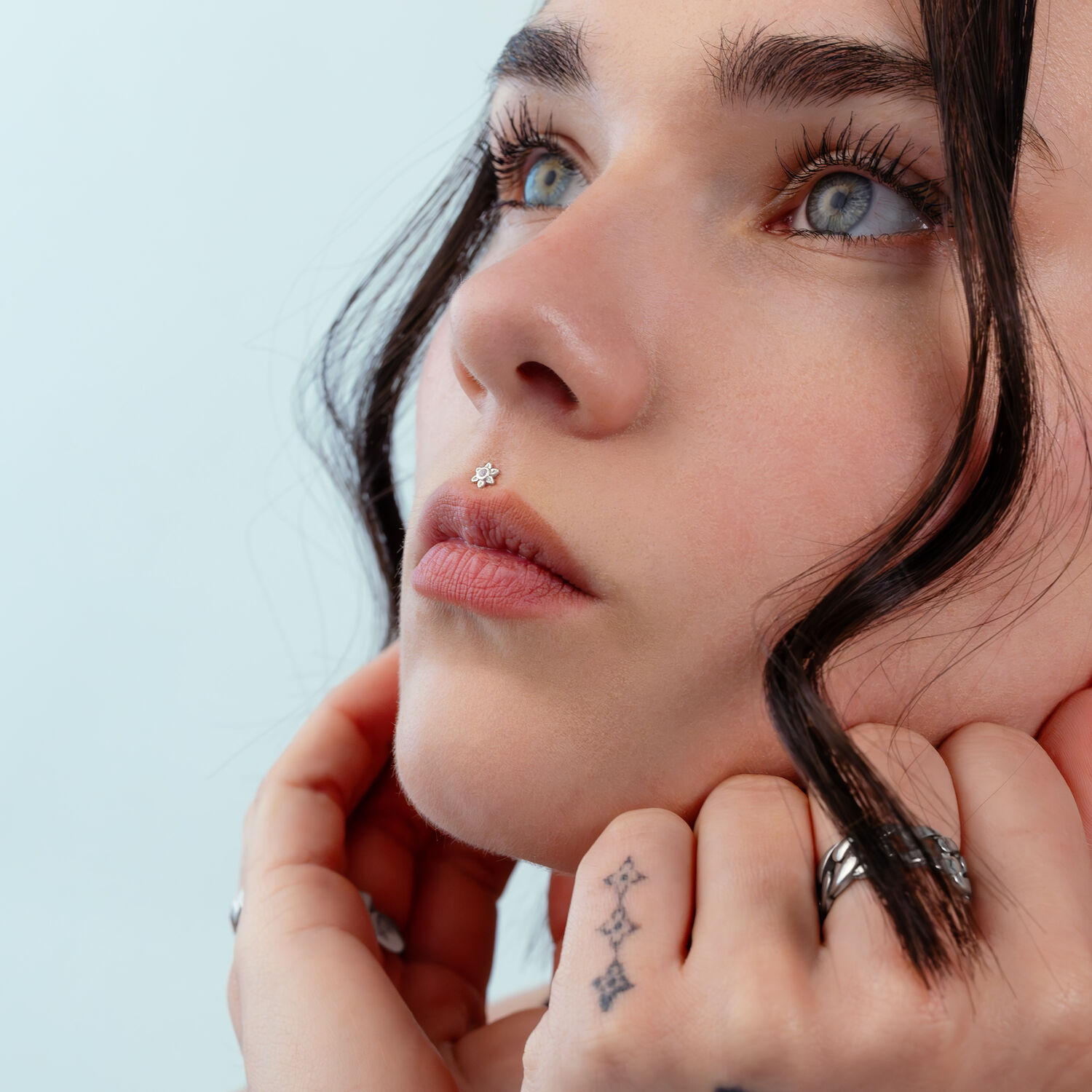 Push Fit Fiore in Argento Primo piano di un viso con un piercing al naso a forma di fiore in argento, sfondo azzurro