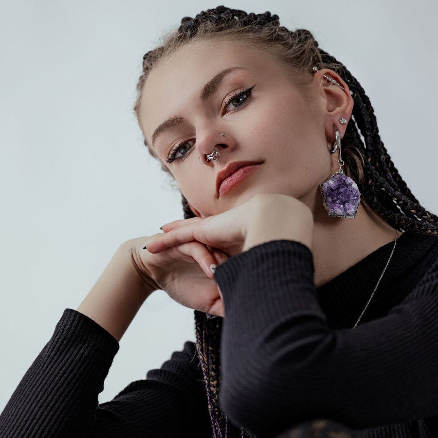 Giovane donna con trecce, orecchino di ametista, piercing al naso, maglione nero su sfondo grigio