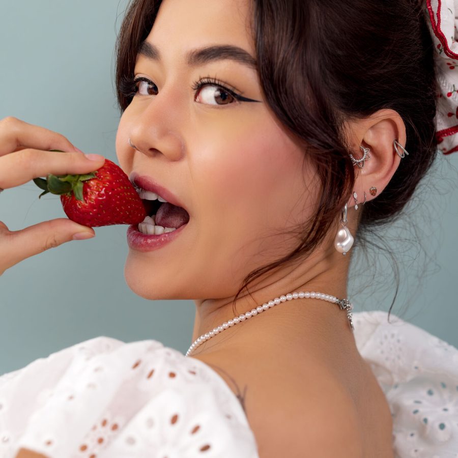 Giovane donna con fragola in mano, diversi orecchini e collana, su sfondo blu