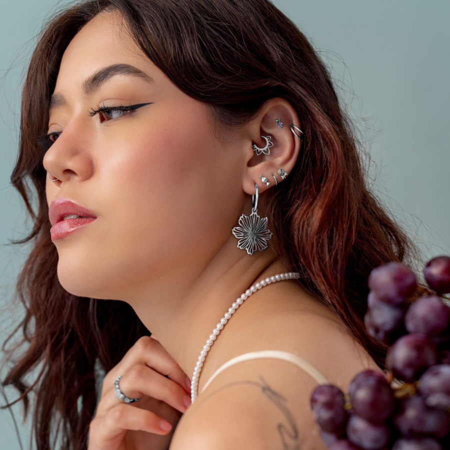 Donna con diversi piercing d'argento all'orecchio, indossa una collana di perle e tiene in mano dell'uva.