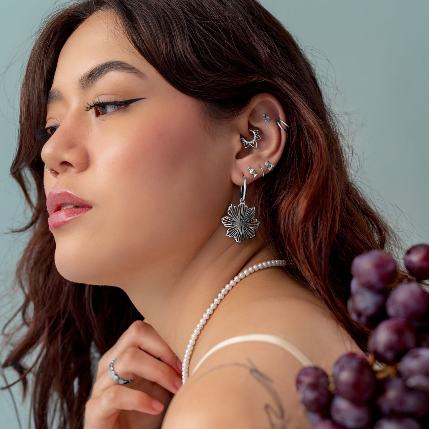 Donna con diversi piercing d'argento all'orecchio, indossa una collana di perle e tiene in mano dell'uva.