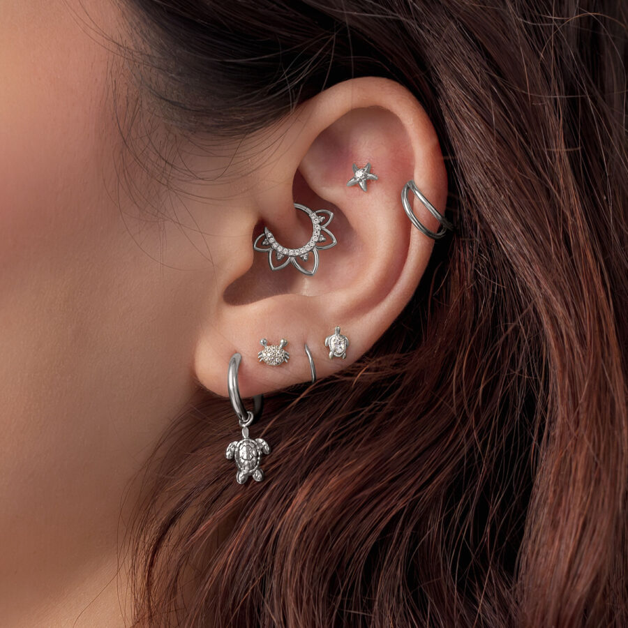 Primo piano di un orecchio con diversi accessori per piercing in argento, incluso un motivo Turtle, su capelli castani.