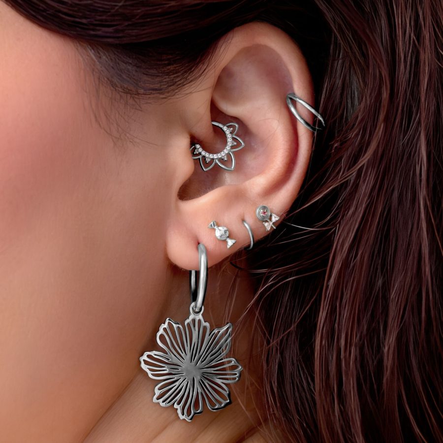 Primo piano di un orecchio con diversi piercing in argento, tra cui un grande ciondolo a fiore e un anello delicato.