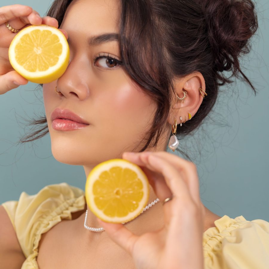 Donna tiene mezza limone davanti all'occhio, indossa piercing dorati alle orecchie, su sfondo blu