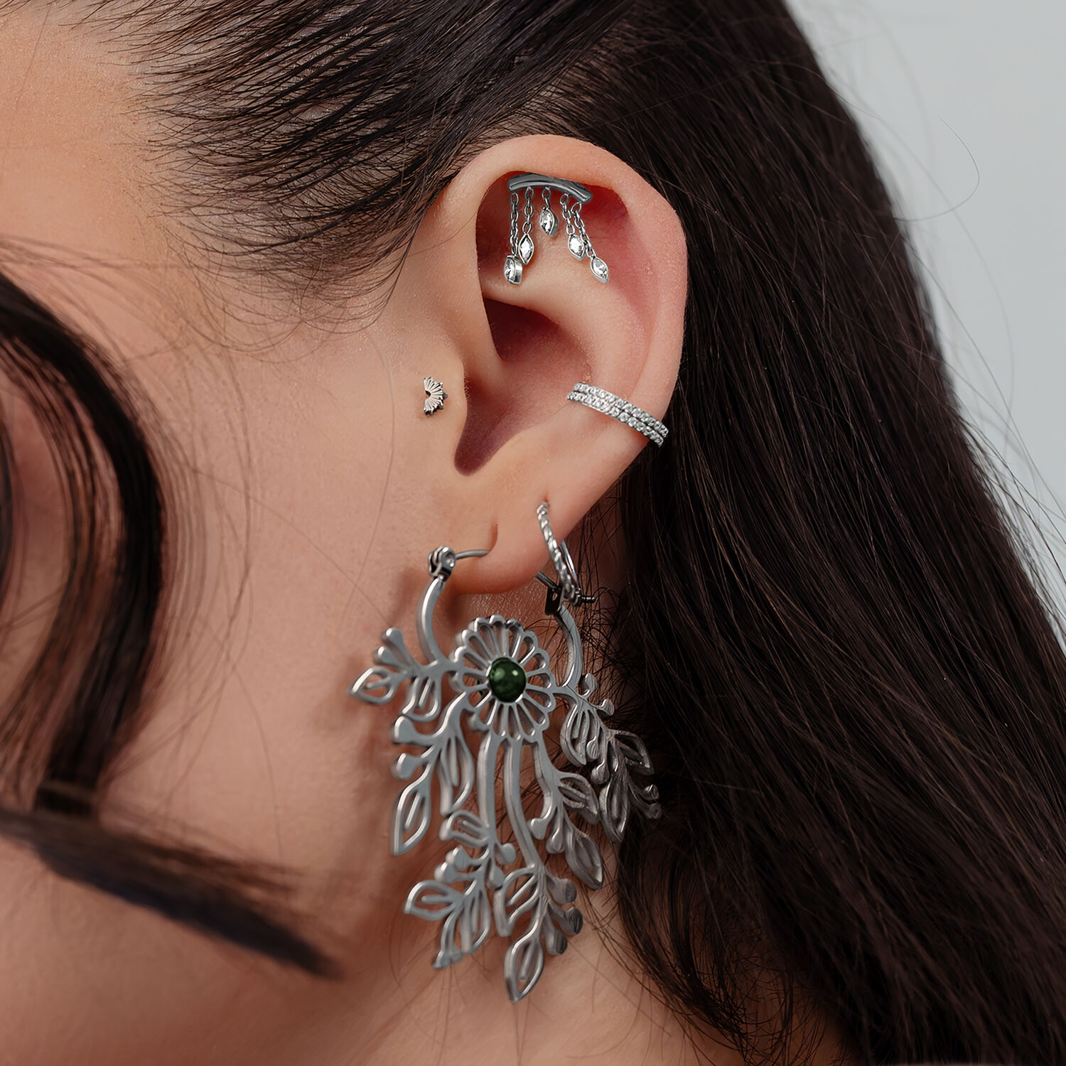 Primo piano di un orecchio con diversi piercing in argento, tra cui un grande pendente ovale e brillanti attacchi a vite.