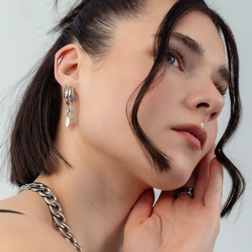 Primo piano di una donna con capelli scuri che indossa orecchini d'argento con catene e una montatura semi-ovale.