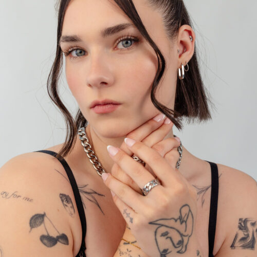 Donna con capelli corti, collana d'argento e diversi anelli, braccia tatuate, sfondo neutro