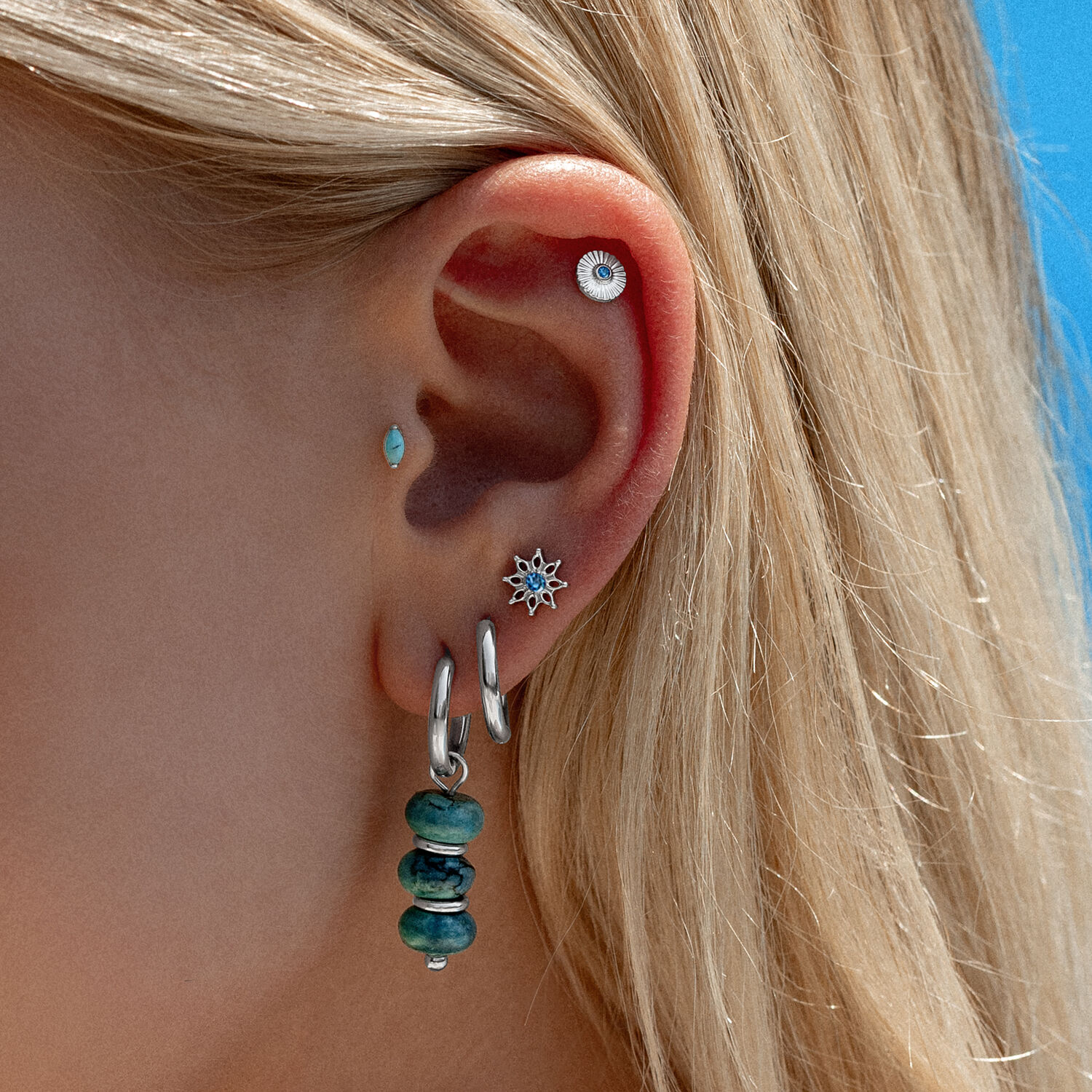 Accessorio per orecchio con cristallo turchese Orecchio con più piercing, incluso accessorio con cristallo turchese e cerchi in argento, davanti a un cielo blu