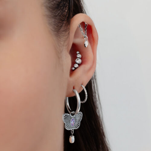 Primo piano di un orecchio con diversi piercing in argento, cristalli e un ciondolo a forma di farfalla su sfondo bianco.