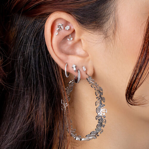 Orecchio con più piercing, tra cui cerchi in argento e inserti di cristallo, capelli castani sullo sfondo