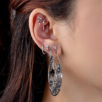 Orecchio con diversi piercing in argento, incluso un grande orecchino rotondo con motivo floreale e cristalli chiari.