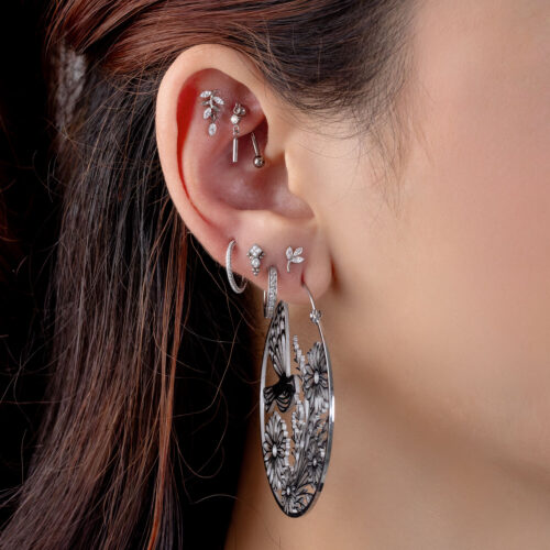 Orecchio con diversi piercing in argento, incluso un grande orecchino rotondo con motivo floreale e cristalli chiari.