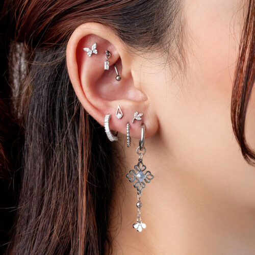 Primo piano di un orecchio con diversi piercing in argento, tra cui un design floreale pendente e cristalli scintillanti.