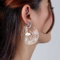 Orecchini a cerchio Summerbird in argento con motivo floreale Orecchini in argento con design floreale e di uccelli, indossati da una donna con capelli castani.