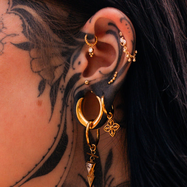 Orecchio con multipli orecchini in oro e tatuaggi neri su sfondo scuro
