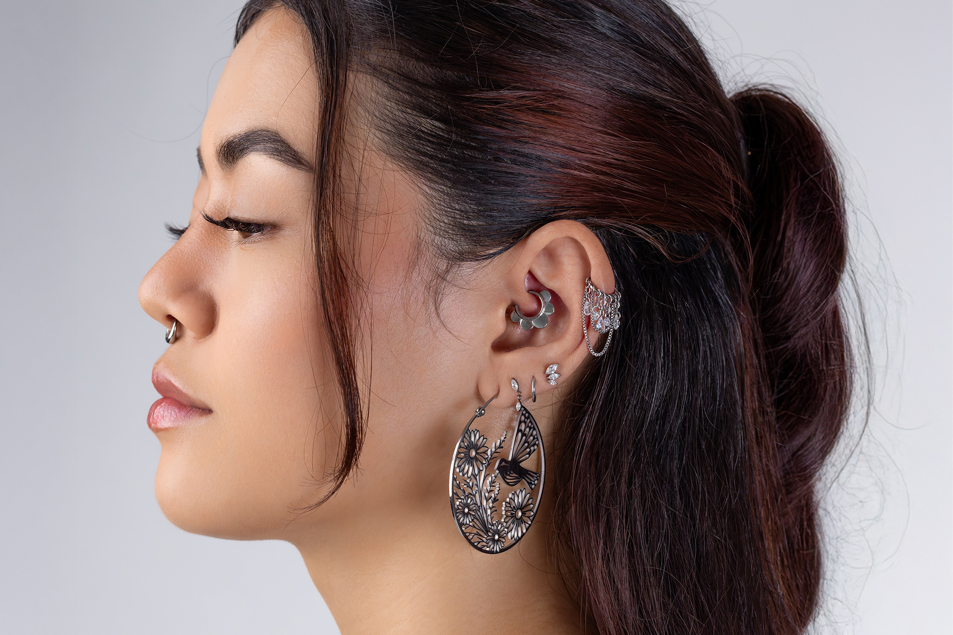 Profilo di donna con capelli scuri, orecchini a cerchio decorati, piercing multipli su sfondo grigio chiaro